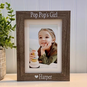 May include: A framed photograph with a gray wooden frame. The text "Pop Pop's Girl" is at the top, and "Harper" with a heart is at the bottom. The photo shows a young girl holding a toy.