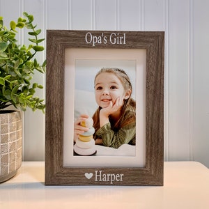 May include: A rustic wood picture frame with a white inscription that reads " Opa's Girl" and a heart symbol followed by the name "Harper". A photo of a young girl is inside the frame.