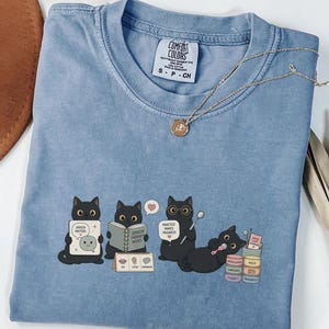 Puede incluir: Una camiseta color azul vaquero con un gráfico de cuatro gatos negros realizando actividades de logopedia. Los gatos aparecen con libros, una tabla de comunicación, un espejo y un sonajero, con el texto Speech Matters y Practice Makes Progress.