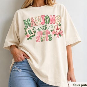 Puede incluir: Una camiseta de manga corta color marfil con un estampado gráfico que incluye el texto "MAHJONG with mah girls". El diseño incorpora patrones de cuadros vichy, flores y cuadros en verde y rosa, junto a ilustraciones de dos fichas de mahjong.