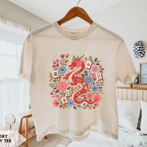 May include: An ivory boxy t-shirt featuring a central graphic of a red dragon surrounded by colorful flowers and various mahjong tiles. The design includes intricate floral details in shades of pink, blue, and green, with mahjong tiles displaying Chinese characters.