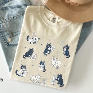 May include: An ivory t-shirt featuring a graphic print of various black and white cats engaged in speech therapy activities, including holding speech cards, playing with blocks, and using a notebook labelled Articulation Practice and Let's Talk.