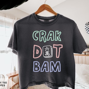 May include: A charcoal grey short-sleeve t-shirt featuring the text CRAK in light green, DOT with a mahjong tile replacing the letter O in light pink, and BAM in light purple. The shirt has a crew neckline and a relaxed, boxy fit.