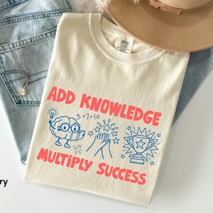 May include: An ivory t-shirt featuring the red text ADD KNOWLEDGE and MULTIPLY SUCCESS. The graphic includes a blue illustration of a brain with glasses reading a book, a math equation, clapping hands with stars, and a trophy with radiating lines.