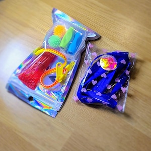 Könnte beinhalten: Zwei transparente Plastiktüten mit verschiedenen Gegenständen. Eine Tüte enthält eine rote Maschenröhre, ein gelbes Reißverschlussarmband und mehrere kleine, bunte Stressbälle. Die andere Tüte enthält einen blauen Gegenstand mit rosa Herzen und einen Aufkleber mit der Aufschrift "Danke".