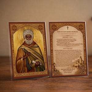 Puede incluir: Dos iconos enmarcados de San Mois&eacute;s el Negro, con detalles dorados e ilustraciones detalladas. El texto en el icono de la derecha describe la vida del santo. Una cruz de madera y una vela encendida est&aacute;n en el fondo, creando un ambiente espiritual.