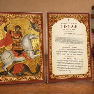Puede incluir: Una placa religiosa rectangular con dos lados: uno muestra un icono de San Jorge sobre un caballo blanco matando a un dragón, y el otro contiene texto titulado Holy Great Martyr George, Victory-Bearer con oraciones.