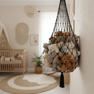 May include: A black macrame net filled with various teddy bears in a nursery setting. The bears are in shades of brown, gray, and white. The net is hanging on a white wall, with a crib and rug visible in the background.