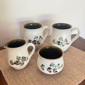 May include: A set of four ceramic items comprising two mugs, one creamer jug, and one sugar bowl. Each piece has a cream-coloured exterior with a dark brown interior and a painted floral motif of small blue, green, and brown leaves.