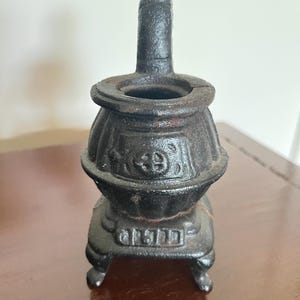 May include: A small, antique cast iron stove with a handle and a round opening. The stove is black with a slightly rusted appearance, and sits on four small feet. The design includes embossed details on the body.