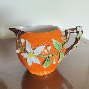 Może przedstawiać: Pomarańczowy ceramiczny dzbanuszek do śmietanki z teksturowaną powierzchnią, zdobiony białymi kwiatami i zielonymi liśćmi. Uchwyt zaprojektowano tak, aby przypominał gałąź. Dzbanuszek ma białe wnętrze i dzióbek.