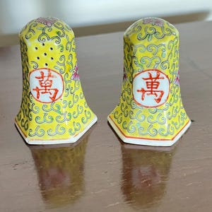 May include: A pair of yellow ceramic salt and pepper shakers with a floral and swirl design. Each shaker has a red and white Chinese character and small holes at the top. The shakers have a hexagonal base.