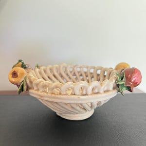 May include: A cream-coloured ceramic bowl featuring a woven lattice design and a pedestal base. The rim is decorated with sculpted, hand-painted fruit accents, including yellow and red pieces with green leaves attached to the sides of the bowl.