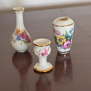 Peut inclure: Trois récipients miniatures en porcelaine avec des corps de couleur crème et des bords dorés. L'ensemble comprend un vase soliflore élancé, une petite coupe en forme de calice et un vase conique, ornés de motifs floraux roses, jaunes et bleus.