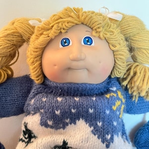 Könnte beinhalten: Eine Puppe mit rundem Gesicht, Grübchen an den Wangen und leuchtend blauen Augen. Sie hat blondes Wollhaar, das zu zwei Zöpfen mit weißen Bändern gebunden ist. Die Puppe trägt einen blau-weißen Strickpullover mit einem winterlichen Baummuster.