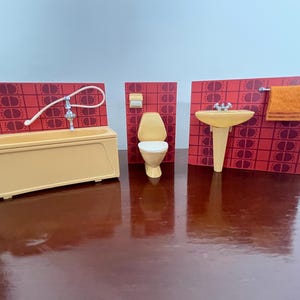 May include: A miniature bathroom set featuring a bathtub, toilet, and sink, all in a light yellow color. The set includes a red patterned backdrop. A small orange towel hangs near the sink. The set is designed for a dollhouse.