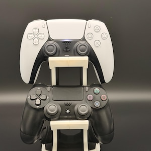 Könnte beinhalten: Ein weiß-schwarzer Gaming-Controller-Ständer, der zwei Controller hält. Der obere Controller ist weiß mit schwarzen Akzenten, und der untere Controller ist schwarz. Der Ständer ist weiß und hat ein modernes Design.