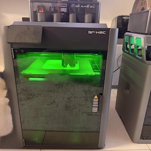 May include: A dark gray 3D printer with a green illuminated interior. The printer has the text "Bambu Lab H2C" on the front. Additional components, including a filament storage unit, are visible above the printer.