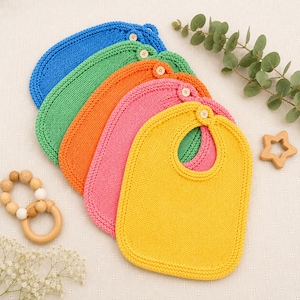 Può includere: Una collezione di cinque bavaglini per bambini lavorati a maglia in colori vivaci: blu, verde, arancione, rosa e giallo. Ogni bavaglino ha una chiusura con bottone e una forma arrotondata. Sono visibili anche una stella di legno e un anello per la dentizione in legno.