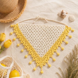 May include: A handmade, cream-colored crochet triangle top with a yellow border and decorative yellow pom-poms. The top is tied at the neck and is displayed on a neutral surface with a straw hat, seashells, and lemons.