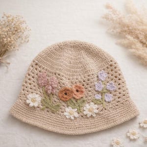 May include: A beige crochet bucket hat adorned with colorful floral embellishments. The flowers include white daisies, pink and purple blooms, and orange and brown accents. The hat has a wide brim and a textured, open weave design.