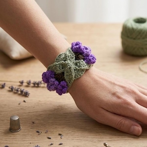 May include: A crochet wrist cuff featuring a sage green braided band accented with clusters of textured purple flowers at each end. The intricate yarn work displays a detailed stitch pattern, creating a floral design that wraps around the wrist.