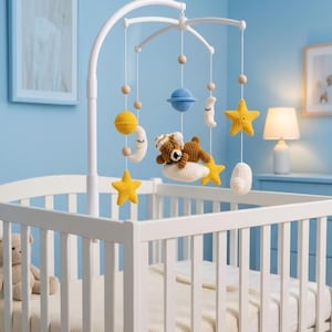 May include: A white crib mobile featuring crocheted figures including a brown bear on a white cloud, yellow stars, white crescent moons, a blue planet, and a yellow sphere, all suspended from a white arched frame with light wooden beads.