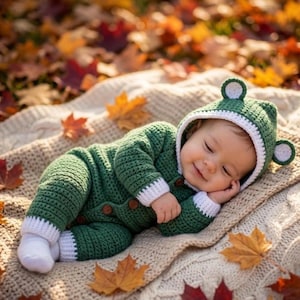 May include: A forest green crocheted baby onesie featuring a hood with two round ears, white trim on the cuffs, ankles, and hood edge, and brown buttons down the front. The garment is crafted with a textured stitch pattern and long sleeves.