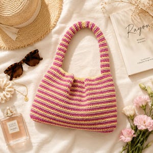 May include: A crochet handbag featuring horizontal stripes in shades of magenta, light pink, and pale yellow. The bag has a single integrated handle and a structured, rectangular body with a soft, textured yarn construction.