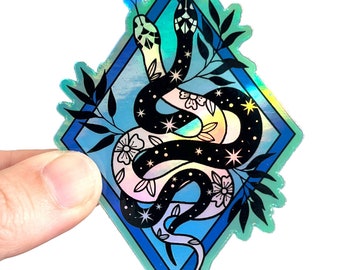 May include: Holographic sticker featuring two intertwined snakes with floral and star details, set within a blue diamond frame. The design includes black, white, and iridescent colors, with leafy accents around the edges. The sticker is held by a hand.