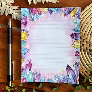 May include: A lined notepad with a watercolor design of colorful crystals and stars. The crystals are in shades of purple, blue, yellow, and pink, framing the lined paper. A silver pen rests on a wooden surface.