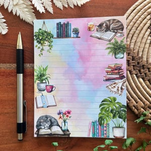 May include: Lined notepad with a watercolor background of pink, blue, and yellow. The notepad features illustrations of books, plants, a cat, and a cup of coffee. A silver and black pen rests on the wooden surface next to the notepad.