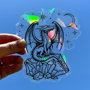 May include: A black and white illustration of a dragon sitting on a pile of crystals. The dragon has wings and a long tail. The background is a holographic sticker with a rainbow effect.