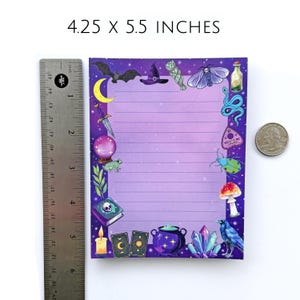 May include: A lined notepad, 4.25 x 5.5 inches, with a purple gradient background and a border of mystical illustrations. The border includes a bat, witch's hat, moon, crystal ball, frog, and other magical elements. A ruler and a quarter are next to the notepad.