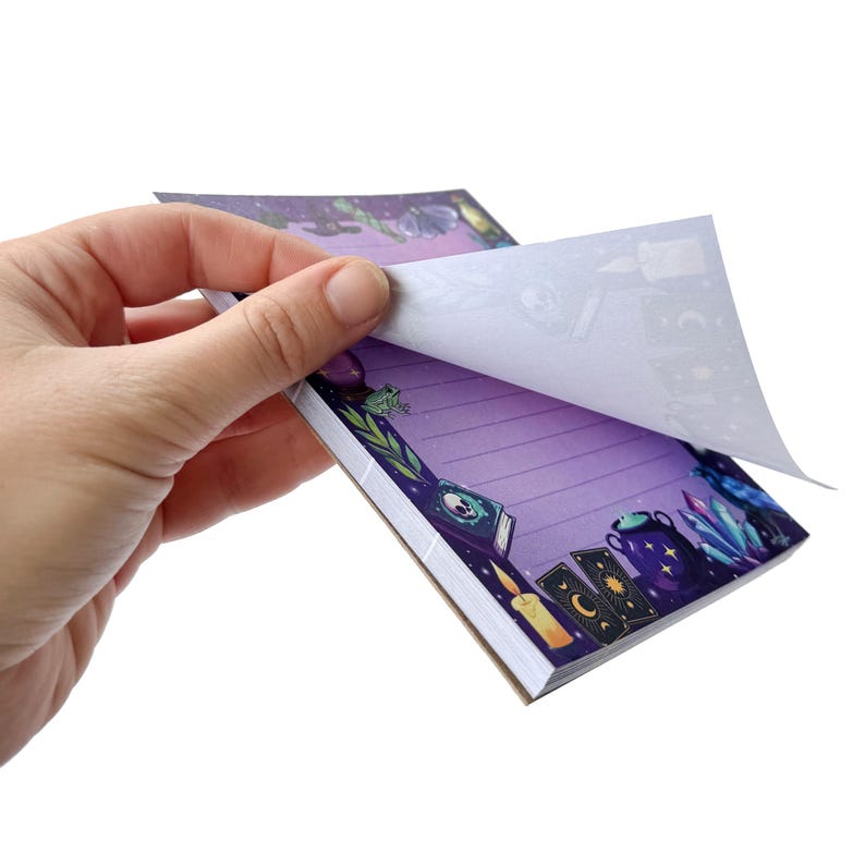 May include: A notepad with a purple background and lined pages. The notepad has a decorative border with illustrations of candles, crystals, books, and tarot cards. The top page is being lifted to reveal the lined pages.