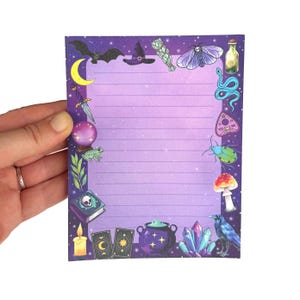 May include: A purple lined notepad with a border of witchy illustrations. The illustrations include a bat, witch hat, sage bundle, moth, bottle, snake, planchette, beetle, mushroom, raven, crystals, cauldron, tarot cards, and a candle.