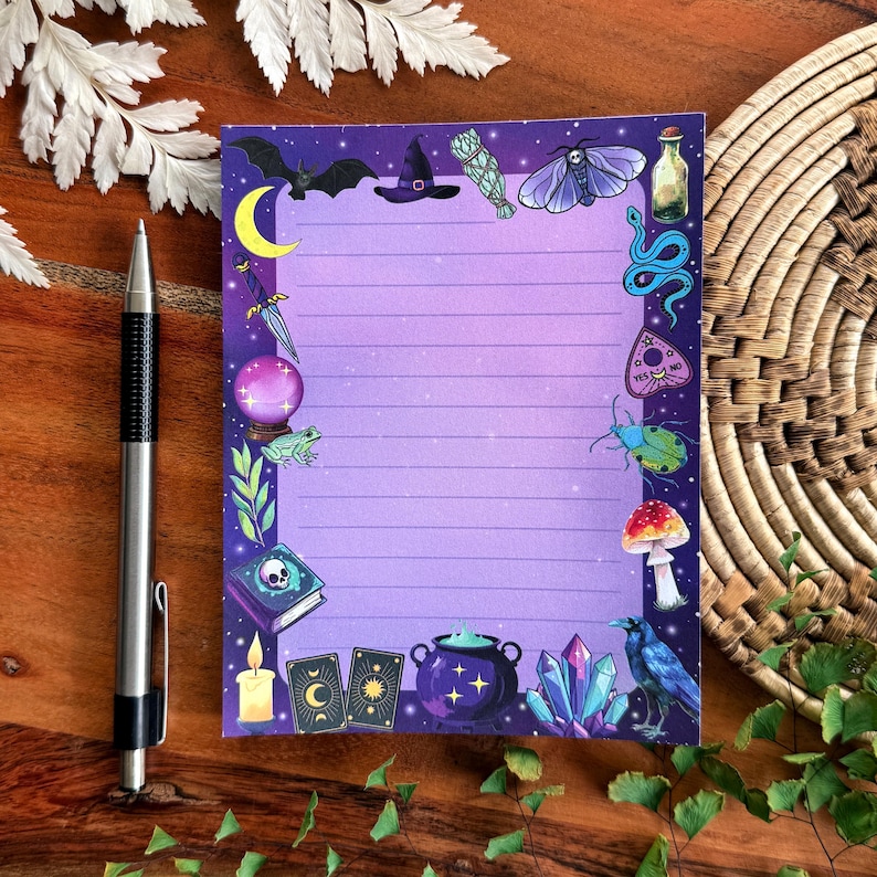 May include: A lined purple notepad with a decorative border featuring witchy and mystical illustrations. The border includes a crescent moon, bat, witch hat, potion bottle, snake, and other magical symbols. A silver pen rests on the wooden surface.
