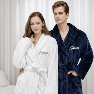 May include: A couple wearing matching plush bathrobes. One is white with "Emma" embroidered on the chest, the other is navy blue with "James" embroidered. Both robes have shawl collars and waist ties.