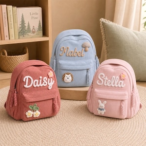 May include: Three corduroy backpacks in pink, light blue, and light pink. The pink bag features the name Daisy and a cherry patch. The blue bag features the name Mabel, a mushroom patch, and a hedgehog patch. The light pink bag features the name Stella and a rabbit patch.