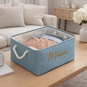 Könnte beinhalten: Hellblaue Aufbewahrungsbox mit weißen Seilgriffen und dem goldfarbenen Namen "Mason". Die Box ist mit gefalteter Kleidung in Rosa und Blau gefüllt. Die Box steht auf einem Holztisch.