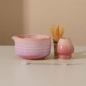 May include: A matcha tea set featuring a pink and purple ceramic bowl with a spout, a bamboo whisk, a pink ceramic whisk holder, and a bamboo scoop. The set is designed for preparing matcha tea.