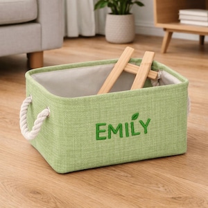 May include: A pale green storage basket with the name "EMILY" embroidered in green. It features rope handles and a light-coloured interior. A wooden item sits inside the basket. The basket is on a wooden floor.