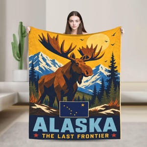 May include: A rectangular throw blanket featuring a large brown moose in the foreground, snow-capped blue mountains, a yellow sun, and dark green pine trees. The Alaska state flag is displayed above the text ALASKA THE LAST FRONTIER in blue.