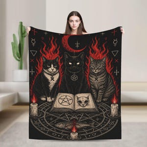 May include: A black throw blanket featuring three cats sitting behind an open book with a pentagram and a skull illustration. The design includes red flames, a crescent moon, occult symbols, a circular geometric pattern, and three lit candles.