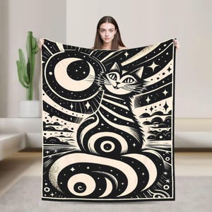 May include: Black and cream-colored throw blanket featuring a whimsical cat design. The cat is depicted with a striped body and a starry background, with a crescent moon and stars. The blanket is held up, showcasing the full design.
