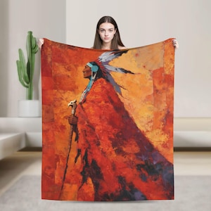 May include: A blanket featuring a Native American figure in profile, wearing a feathered headdress and a red cloak. The artwork has a warm color palette of reds, oranges, and yellows, with a staff in hand. The blanket is held up by a person.