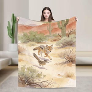 May include: A rectangular throw blanket featuring a watercolor illustration of a desert landscape. The scene depicts a coyote and a roadrunner moving across sandy terrain with sagebrush, cacti, and distant orange-toned mesas in the background.