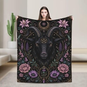 May include: A black rectangular throw blanket featuring a central illustration of a goat head with a pentagram on its forehead. The design includes a crescent moon, purple and pink floral accents, and intricate gold filigree patterns throughout.