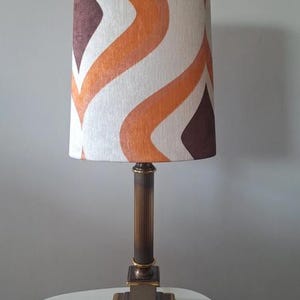 Op de afbeelding: Een tafellamp met een cilindrische lampenkap voorzien van een golvend, herhalend patroon in oranje, bruine en crèmekleurige tinten. De lampvoet is gemaakt van donker metaal met een gecanneleerde kolom en een vierkante, getrapte voet.