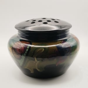 May include: A ceramic flower frog bowl with a glossy black lid featuring multiple circular perforations. The rounded base displays a dark, multi-colored abstract pattern in shades of deep green, brown, and red with subtle gold-toned outlines.